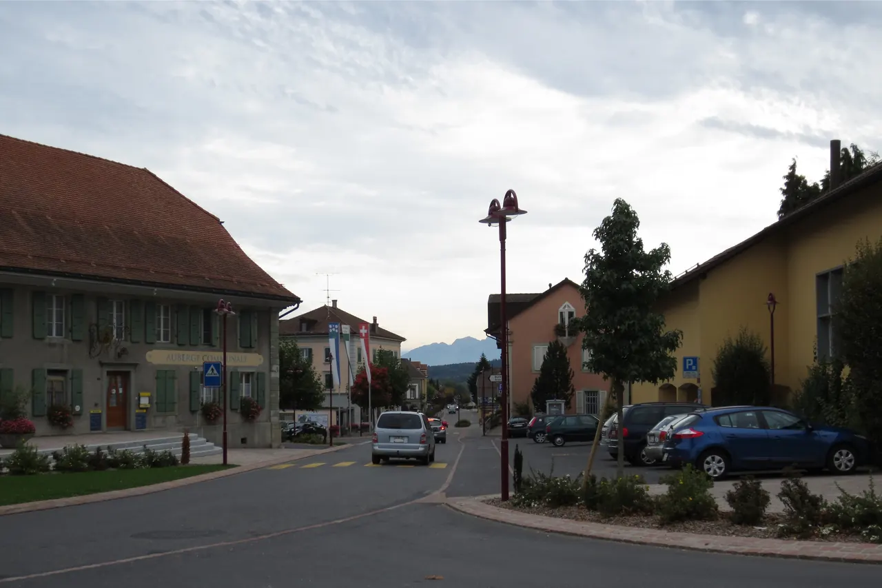 Rue principale de Mézières : grande ferme à toiture rouge, maison jaune et voitures